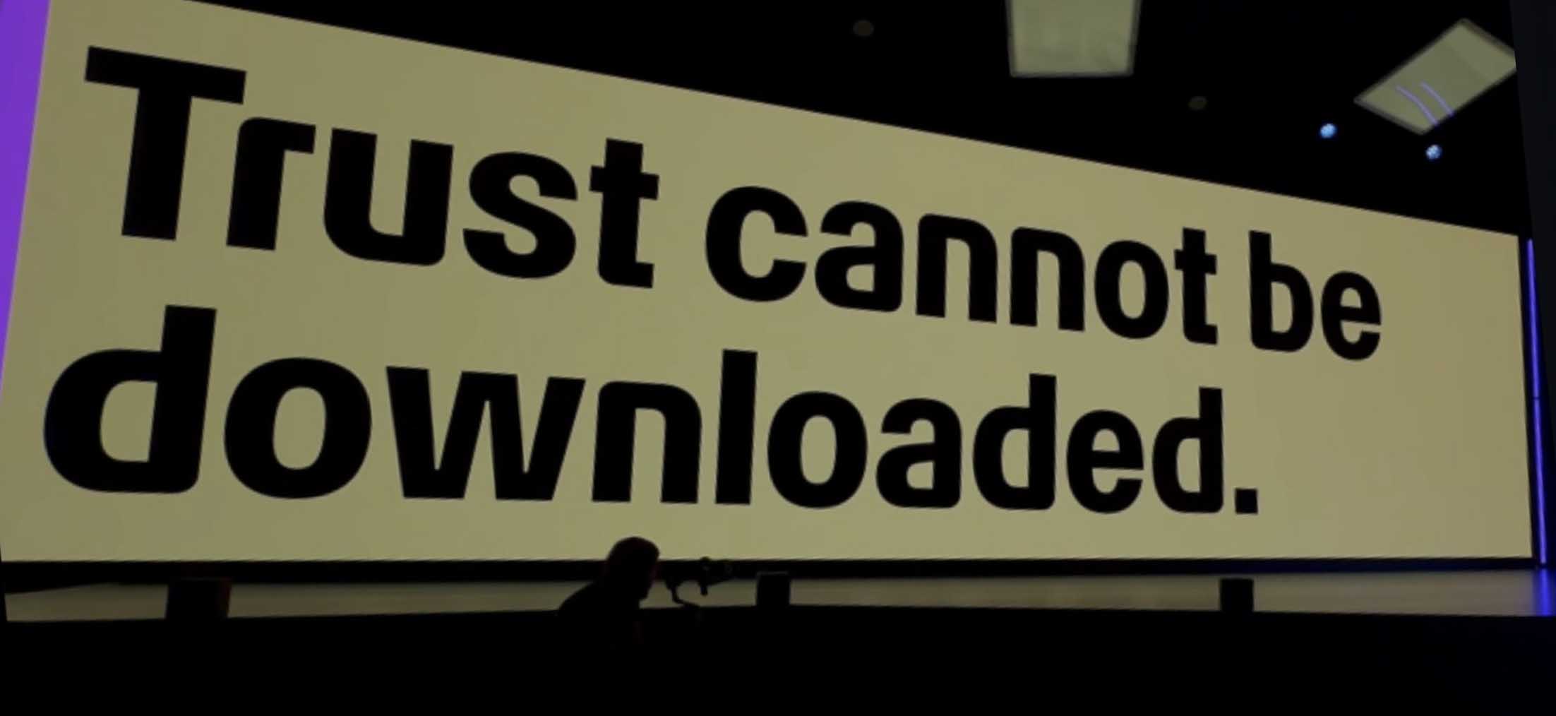 Bühnenprojektion mit dem großformatigen Schriftzug Trust cannot be downloaded auf gelbem Hintergrund, der Sprecher steht klein rechts daneben vor einer zweiten Leinwand mit dem IFA-Logo