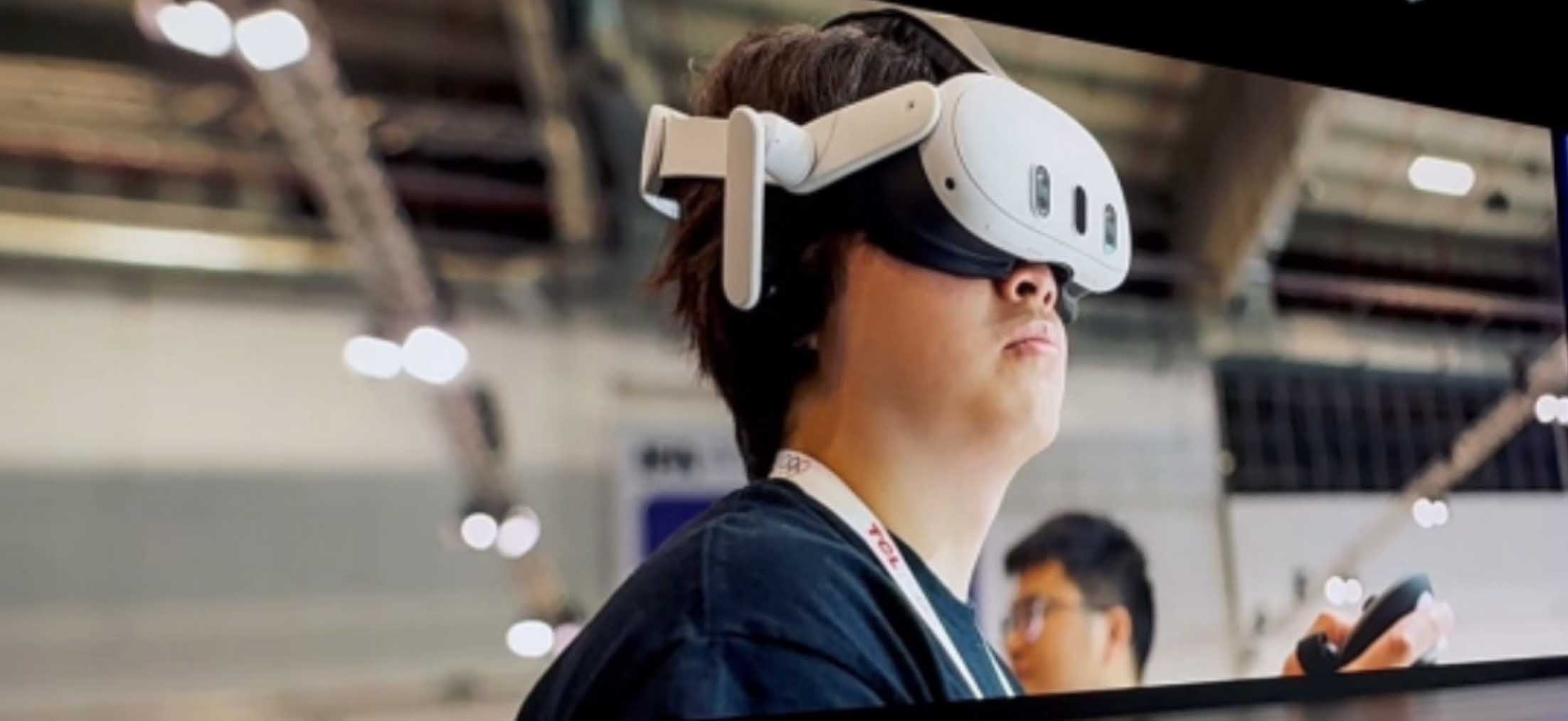 Großformatige Bühnenleinwand mit dem Live-Foto eines jungen Messebesuchers, der ein weißes Mixed-Reality-Headset mit Controllern trägt, aufgenommen vor dem Hintergrund einer Ausstellungshalle, rechts daneben der Sprecher klein vor einer gelben Leinwand mit IFA-Logo